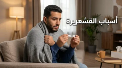 رجل عربي شاب يجلس على أريكة ملفوفاً ببطانية خفيفة ويمسك ميزان حرارة رقمي مع ظهور عبارة أسباب القشعريرة
