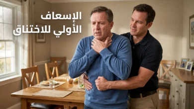 رجل يُطبّق مناورة هيمليك على شخص بالغ يختنق في غرفة طعام منزلية، مع توضيح وضعية اليدين الصحيحة فوق السرّة مباشرة — الإسعاف الأولي للاختناق