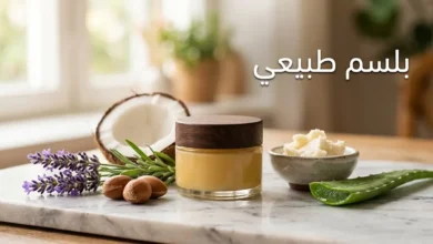 برطمان زجاجي أنيق يحتوي على بلسم طبيعي ذهبي محاط بمكس أرغان وجوز هند ولافندر وزبدة شيا وألوفيرا على سطح رخامي بإضاءة طبيعية